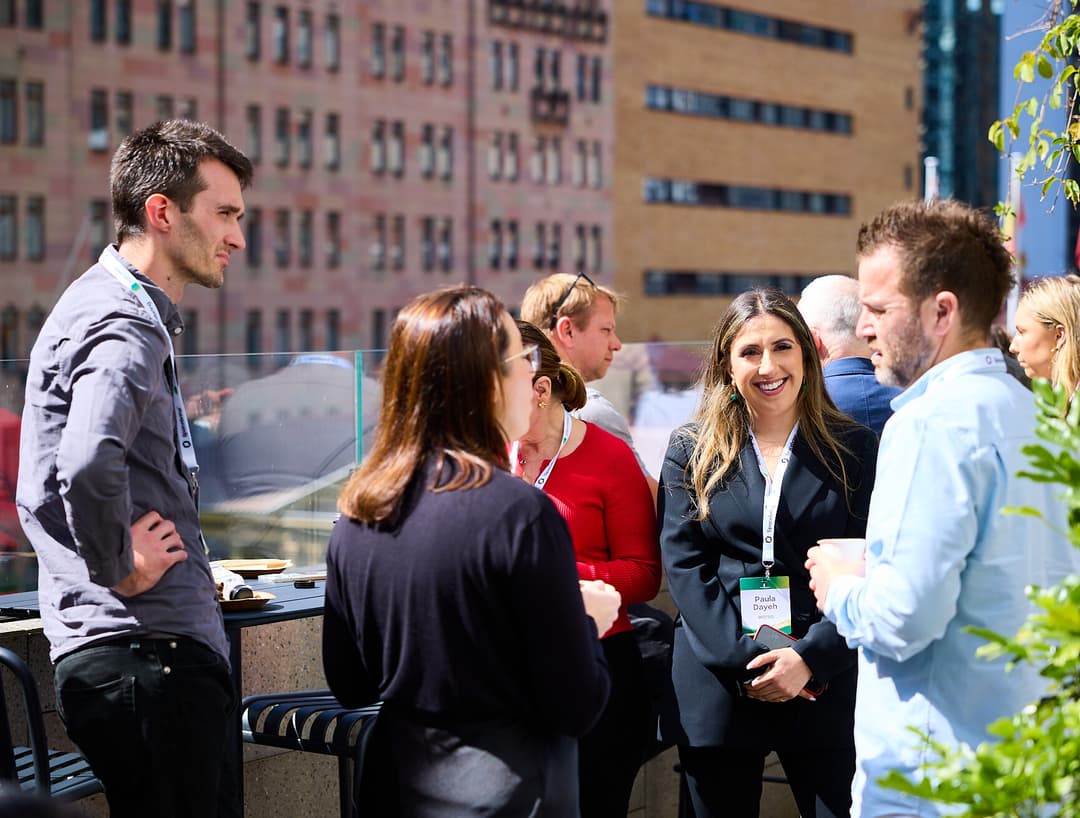 A group of people socialize and network outdoors on a sunny rooftop, wearing conference badges. City buildings are visible in the background, and greenery frames the scene. The Flexible Workspace SF logo is in the corner.