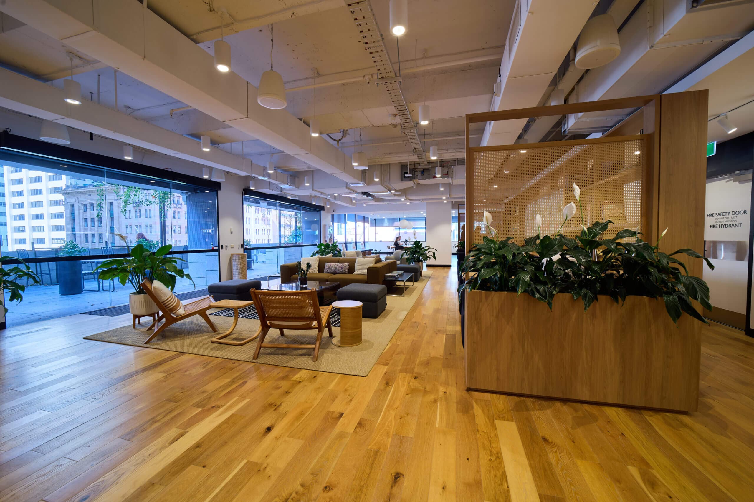 A brightly lit, modern office lounge area with hardwood floors. The space features couches and chairs arranged on area rugs, a wooden planter box filled with peace lilies, and large windows offering a view of a city building. Natural light floods the space creating a relaxed atmosphere.