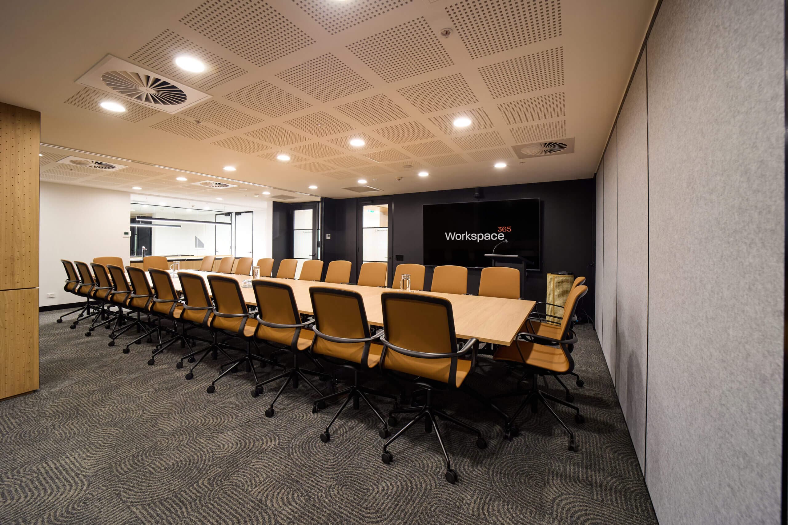 An office conference room is set up with a long, light wood conference table surrounded by multiple tan mesh-backed office chairs. A large dark flat screen is mounted on the rear wall. The carpet is grey with a swirling design. The ceiling is off white with inset lights and vents.