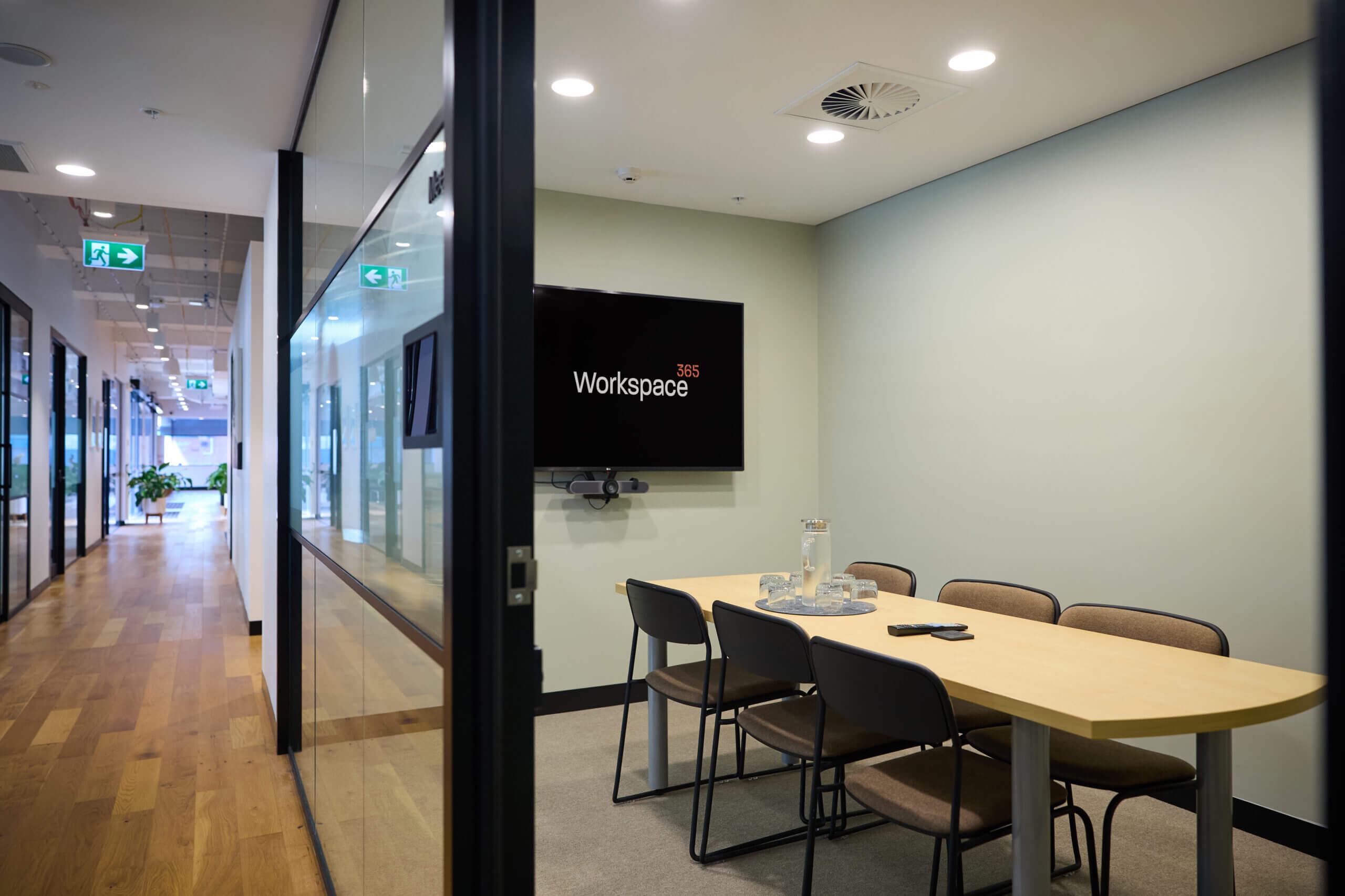 Wide-angle, indoor shot. A meeting space with a rectangular light wood table and 6 brown chairs. The wall has "Workspace 365" displayed against a dark screen, with a video conferencing camera mounted below. From the hallway, wood flooring with emergency exit lighting. The foreground features a dark frame of the meeting room, creating depth, with black framed glass walls. Well-lit, muted color palette.