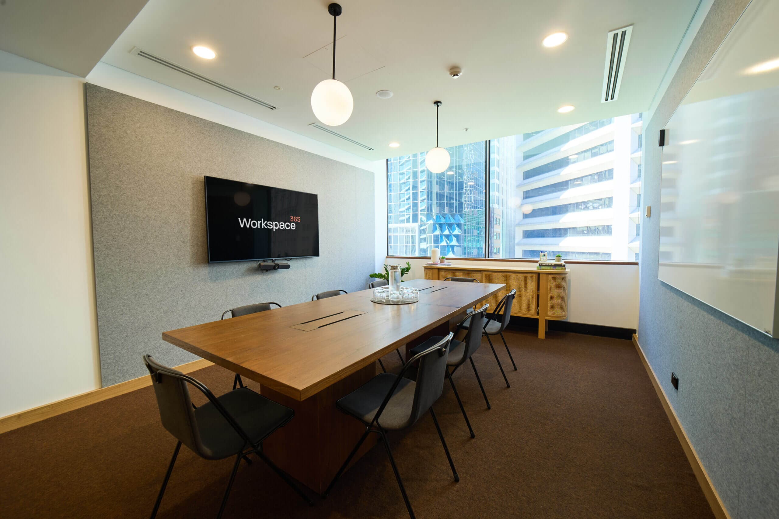 Interior shot of a well-lit conference room. The composition is centered on a long, rectangular wooden conference table with black chairs. A dark brown carpet covers the floor. On the left, a gray acoustic panel wall mounts a black flatscreen monitor displaying "Workspace 365". A large, floor-to-ceiling window on the right reveals a cityscape of closely packed office buildings. Two round pendant lights with a matte finish hang above the table. The room is lit with recessed lighting to cast soft, even light.