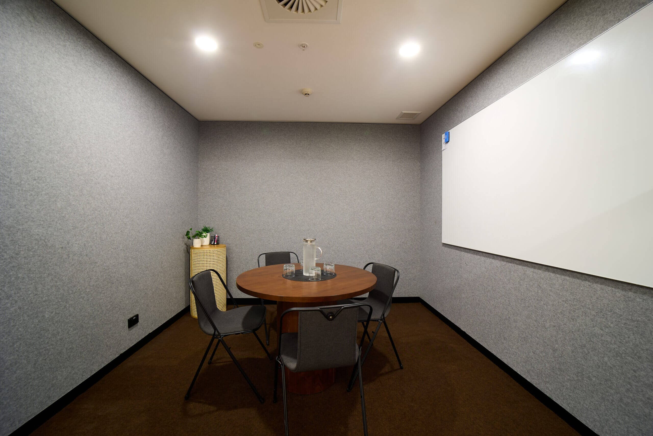 Small meeting room with round table, four chairs, whiteboard, and gray felt walls.