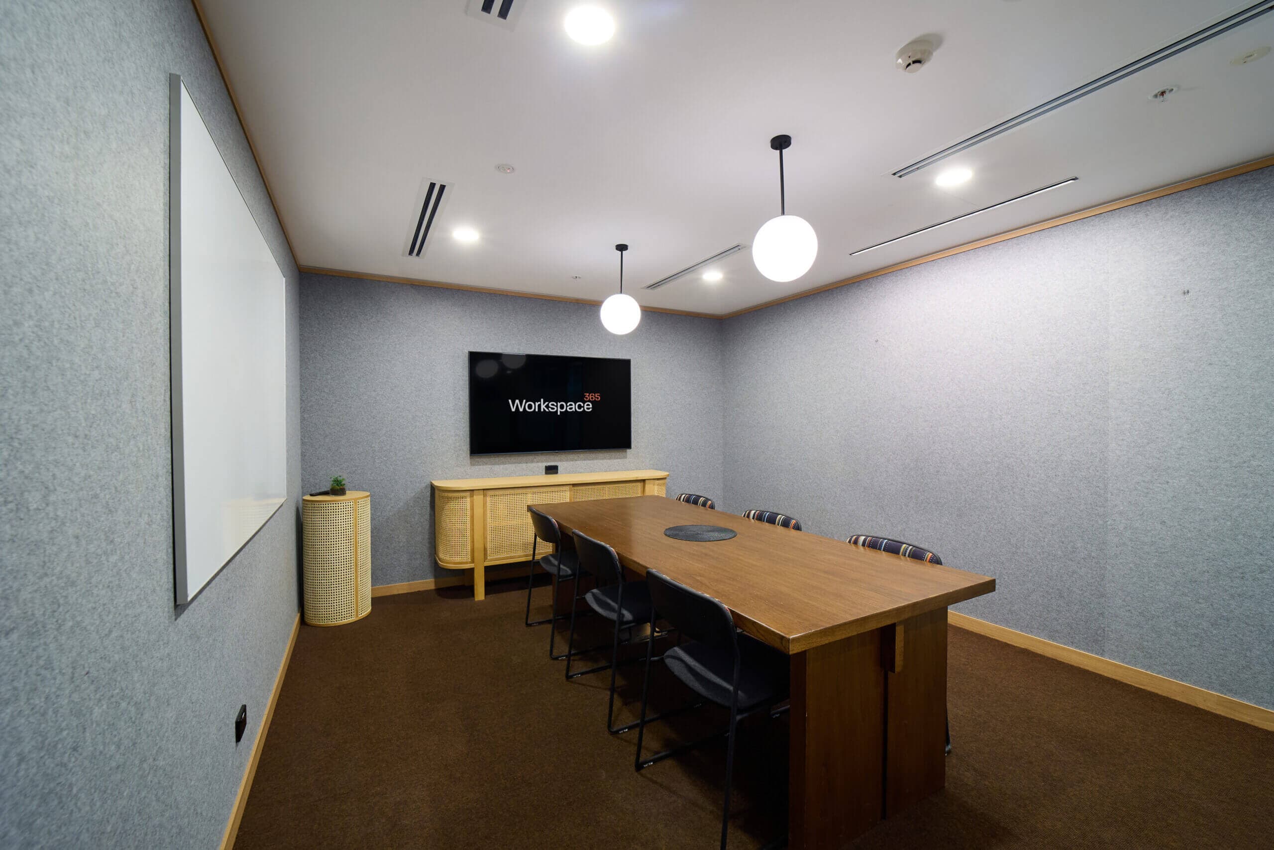 A well-lit, small conference room features a wooden table surrounded by black chairs. A large screen and a whiteboard are mounted on the wall. The room has soft, gray, sound-dampening textured walls and minimalist décor.