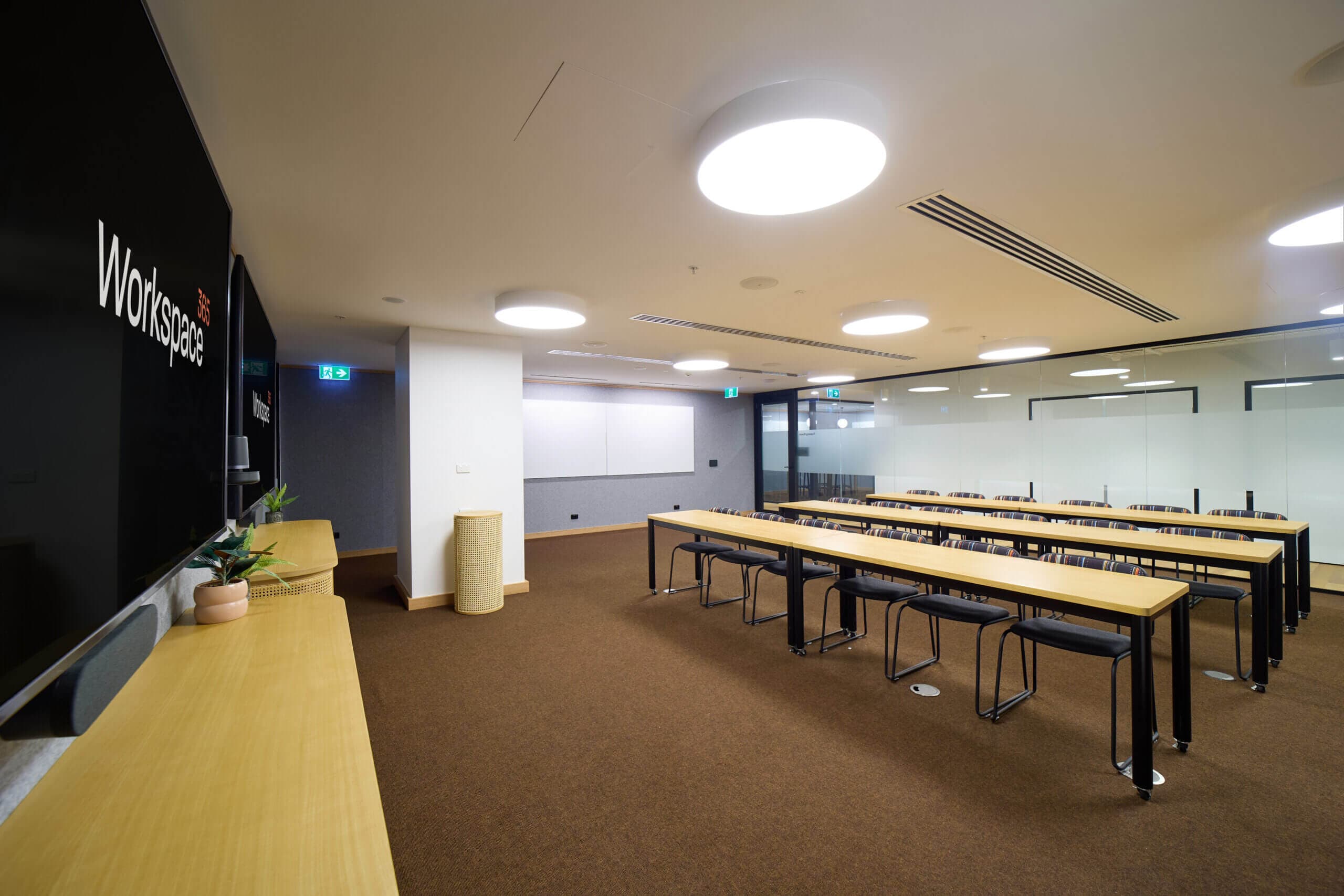 Interior shot of a contemporary training room. Tables with black chair are set up in rows. The walls are light gray, with a large visual presentation equipment. Round ceiling lights provide illumination. The flooring is brown carpet. The image is composed with a slight wide-angle lens effect, showcasing the long perspective of the room.