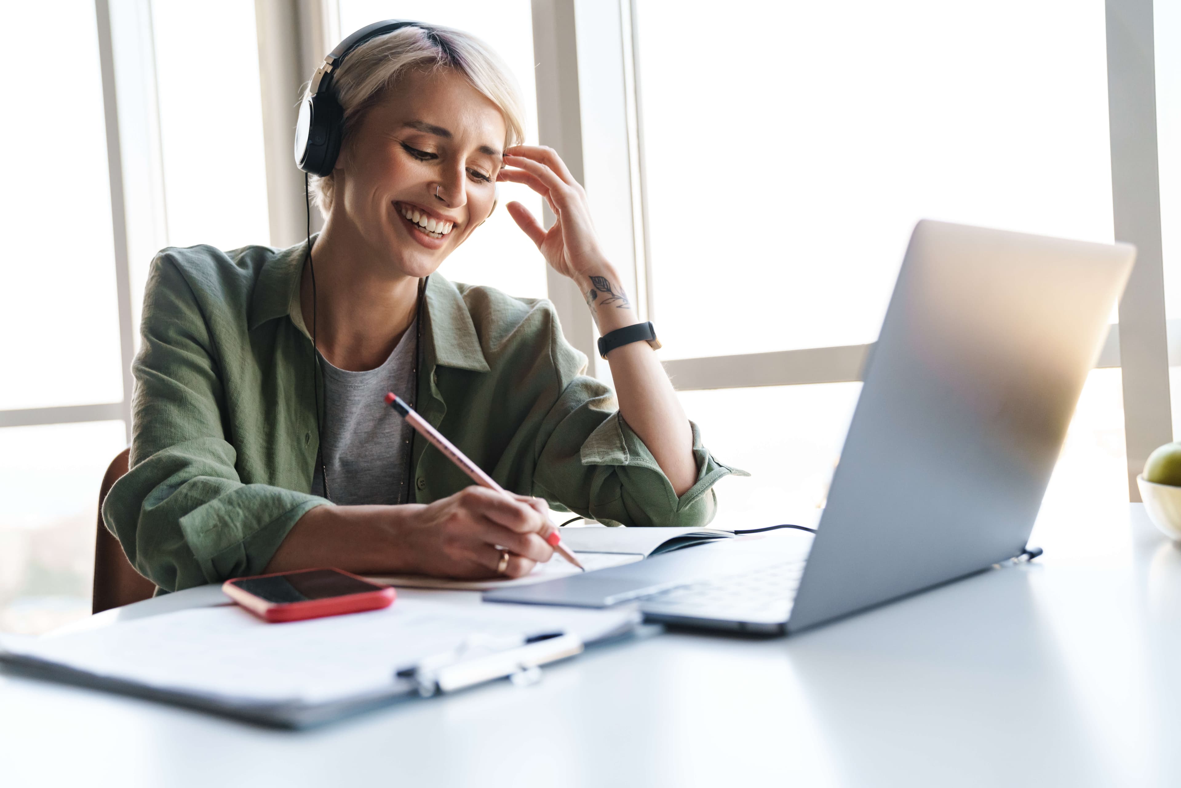A smiling woman wearing headphones sits at a desk, writing in a notebook while looking at her laptop. A smartphone, clipboard, and apple are also on the desk by a bright window.