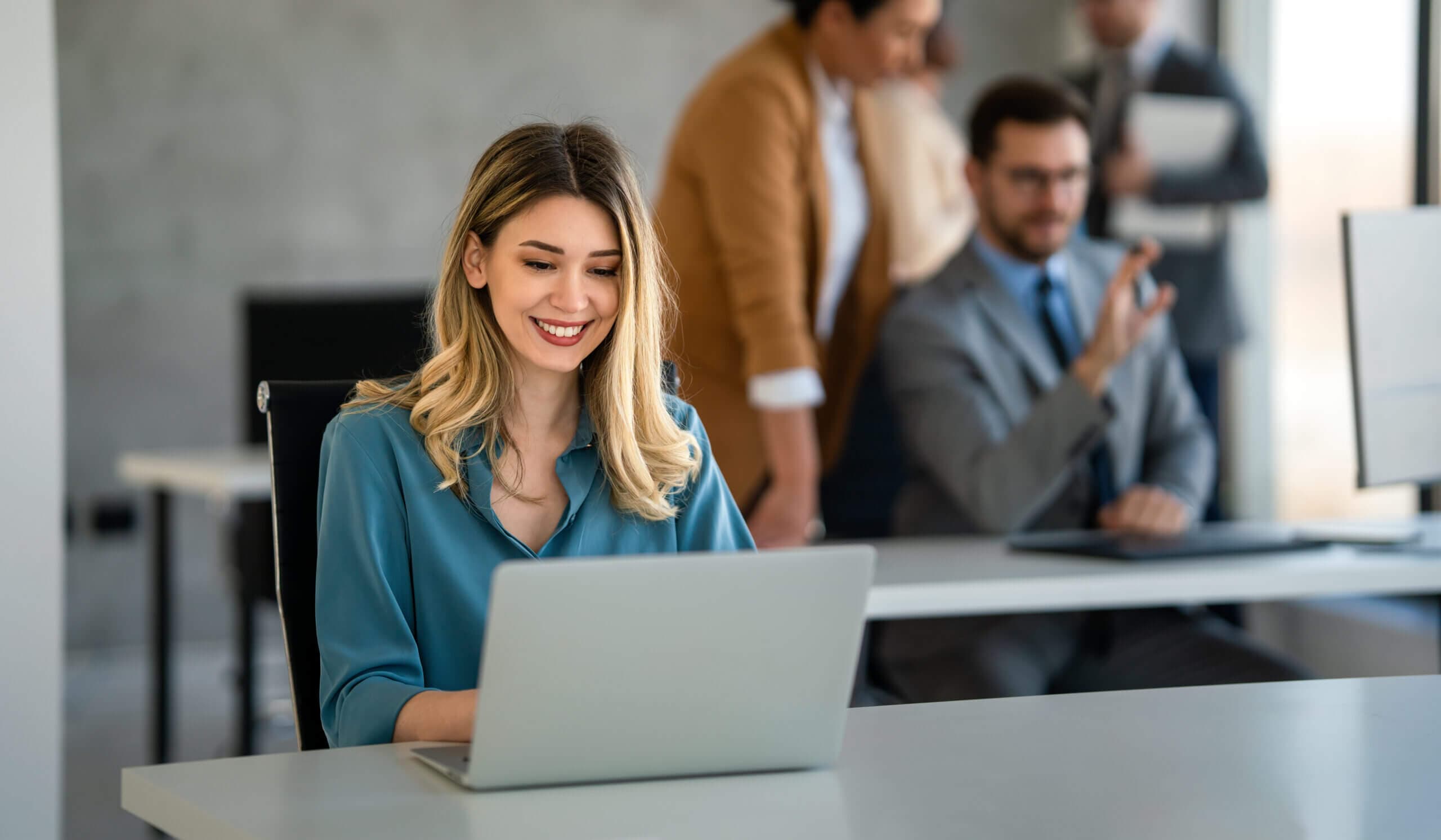 A woman with long blonde hair and a teal blouse is smiling at her laptop in an office. In the background, a man in a suit with glasses gestures while talking to someone. Two more people are engaged in conversation near them.