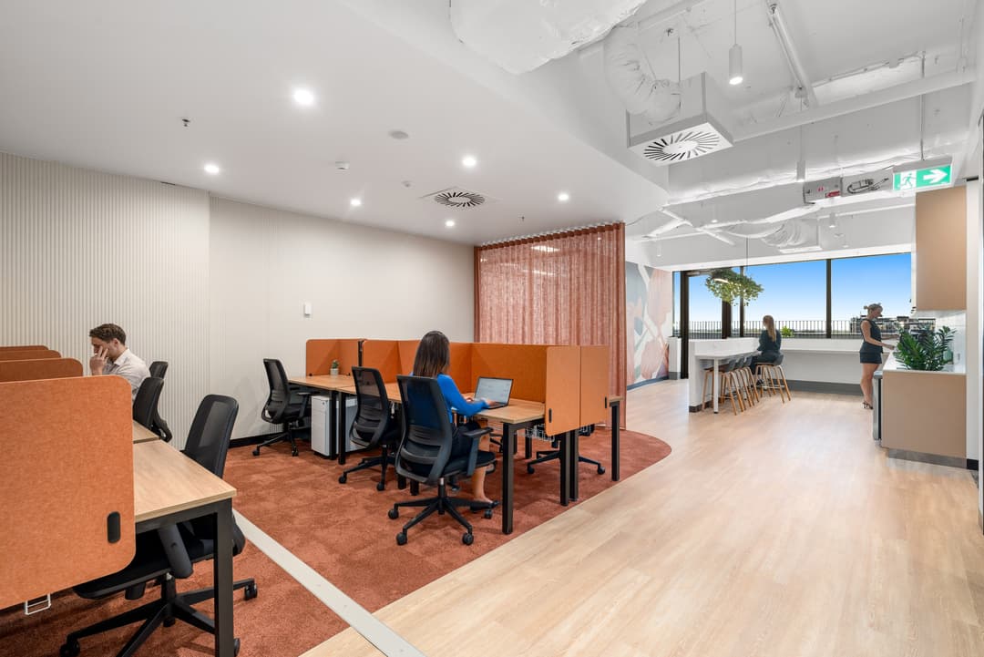 Modern office with individual workstations and orange dividers, people working on laptops, bar seating by large windows, and a bright, open atmosphere with wood floors and ceiling lights.